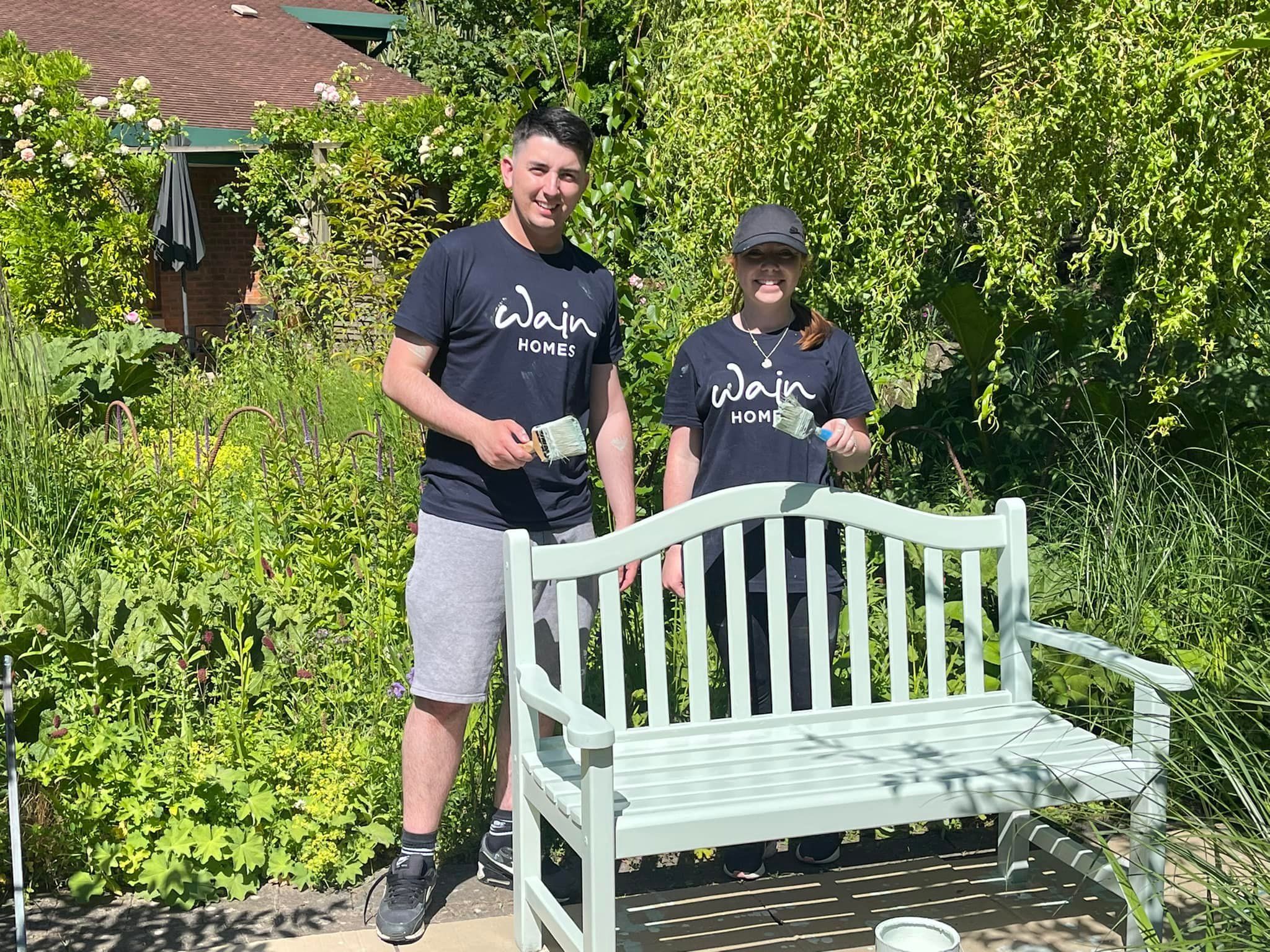 A man and a woman are standing in black T-shirts with the Wain Homes logo on behind a bench they have painted in pale green